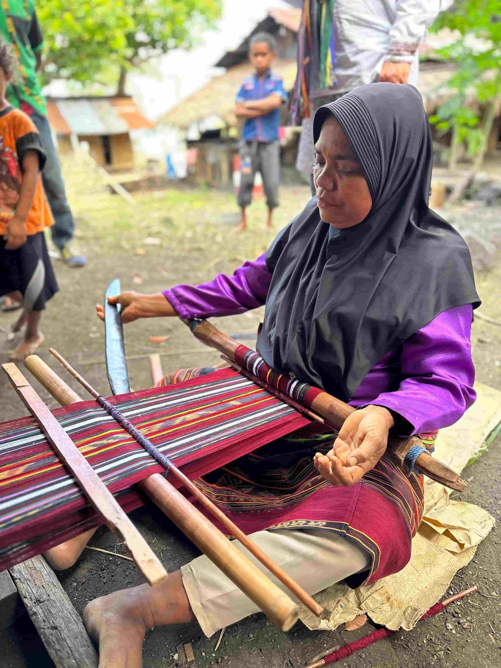A woman in a purple shirt and hijab weaving an ikat cloth on a backstrap loom